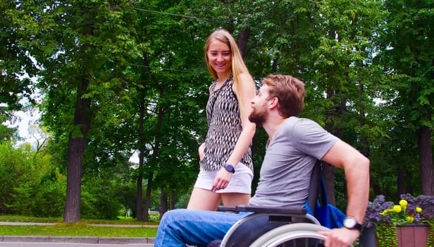 Happy young disable man in wheelchair walking in the park with his wife. Side view.