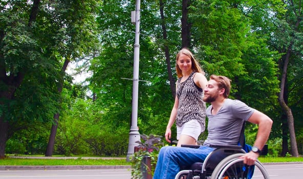 Happy young disable man in wheelchair walking in the park with his wife. Side view.