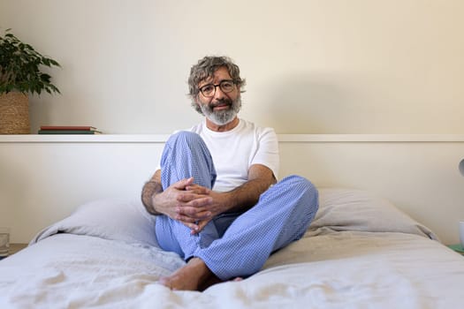 Mature man wearing pyjamas relaxing sitting on the bed looking a camera. Lifestyle concept.