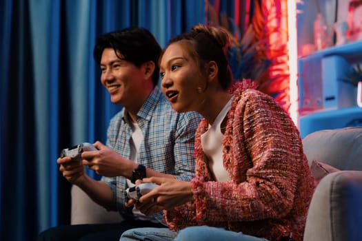 Sitting sofa of happy smiling couple playing video game control by joystick fronted snack and drinks competing challenge level with funny at modern neon blub light comfy living room at home. Infobahn.