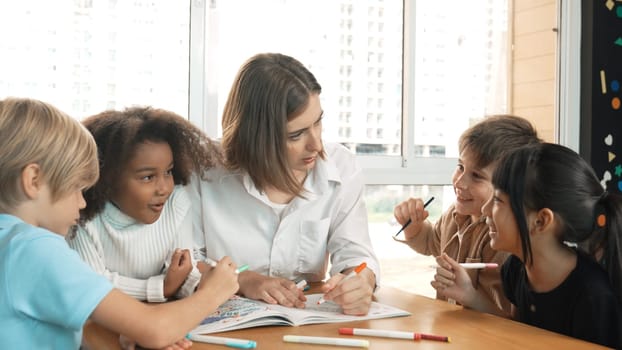 Professional caucasian teacher telling story to diverse student while sitting at table with storybook and colored book. Smart learner listening story while colored picture from instructor. Erudition.