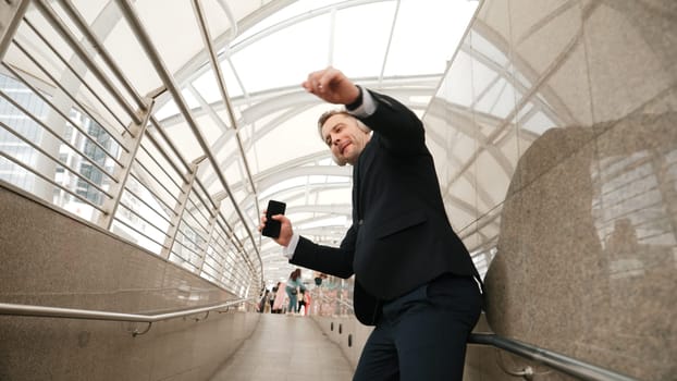 Business man dance to relaxing music from headphone at modern corridor. Cheerful project manager wear headset and moving gesture to celebrate successful song with lively mood in urban city. Urbane.