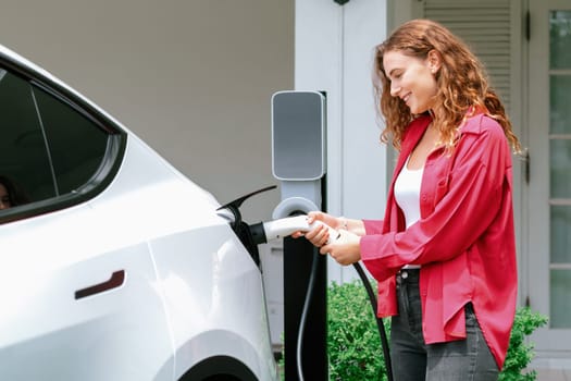 Modern eco-friendly woman recharging electric vehicle from home EV charging station. EV car technology utilized for home resident to future environmental sustainability. Synchronos