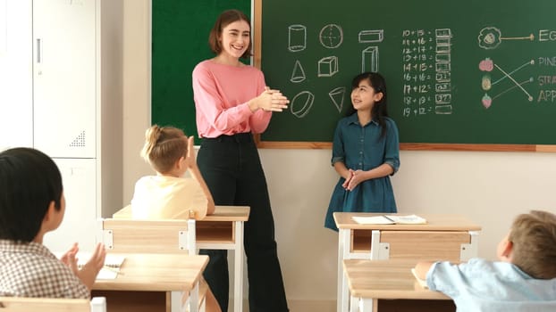 Caucasian teacher asking for volunteer while asian girl walking at in front of class with confident. Smart student raised hand and answer question while teacher clapping hands to encourage. Pedagogy.
