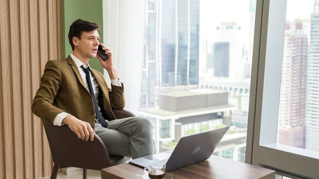 Businessman sitting on furniture working on laptop at ornamented corporate waiting area with cityscape background on the window. Business profession and strategic marketing plan for business success.