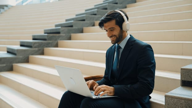 Professional business man sitting at stairs while working on laptop. Skilled project manager listening music from headphone and checking email and discussion about marketing plan. Outdoor. Exultant.