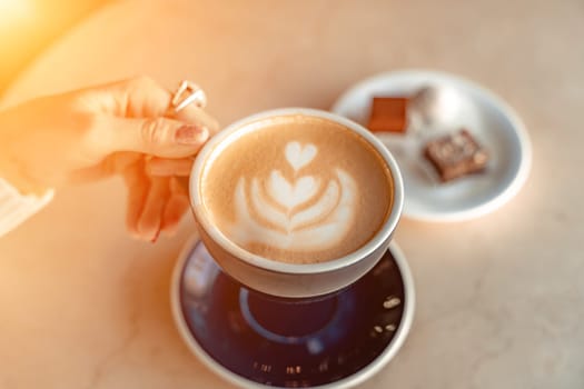 A cup of fresh cappuccino coffee in the hands of a woman on a fashionable background of a white marble table, next to a plate with sweets. Coffee addiction. Top view, flat lay