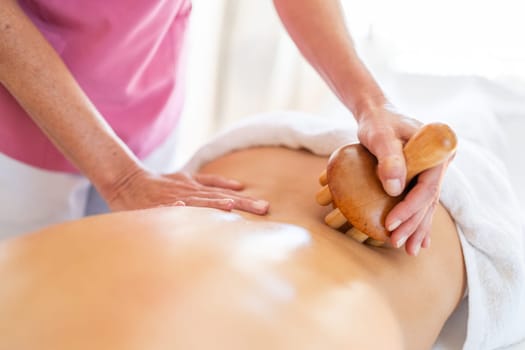 From above of crop anonymous massage therapist massaging with wooden therapy tool back of client lying on table during osteopathy session in clinic