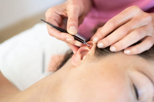 From above crop homeopath professional beautician applying auriculotherapy ear acupuncture techniques on woman client in modern clinic