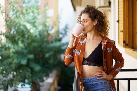 Young stylish woman wearing trendy outfit and drinking coffee while standing on balcony of modern residential house against blurred background