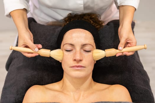 Crop beautician use brushes to apply facial cream on face of young female customer in beauty centers during cosmetic session and skin care