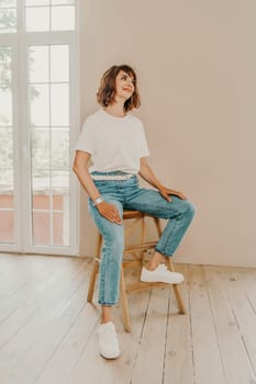 Portrait of brunette white t-shirt. A woman sits on a chair and looks at the camera, business portrait on a beige background, dressed in a white t-shirt and jeans