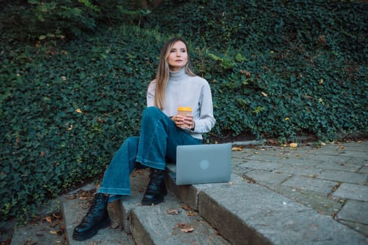 A woman sits on the ground with a cup in her hand. She is wearing a gray sweater and blue jeans