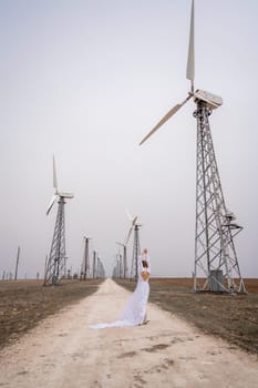 A woman in a white dress is walking down a dirt road in front of a row of wind turbines