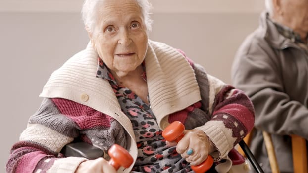 An aged old woman exercising with dumbbells sitting on a nursing home