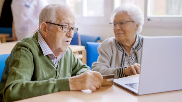 Senior woman explaining how to shop online to her husband using laptop and credit card