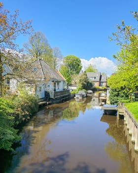 A winding canal river gracefully flows through a vibrant green landscape, surrounded by lush trees, grassy meadows, and rolling hills under a clear blue sky. Broek in Waterland village Netherlands