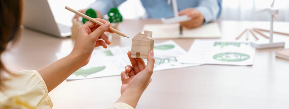 Cropped image of businesswoman presents eco-friendly house by using green design to manager while holding the wooden house block at table with windmill model placed with document. Delineation.