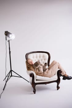 Female blogger, blonde on chair with domestic cat scottish straight in white photo studio. Dressed in a formal beige trouser suit. Vertical