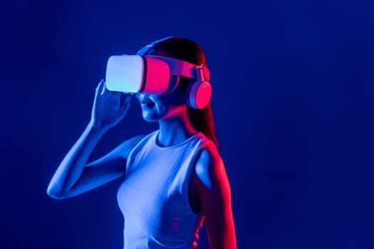 Smart female stand with surrounded by cyberpunk neon light wear VR headset connecting metaverse, futuristic cyberspace community technology. Elegant woman look generated virtual area. Hallucination.