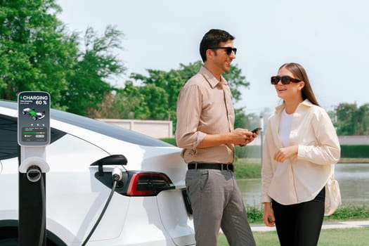 Young couple use smartphone to pay for electricity at public EV car charging station green city park. Modern environmental and sustainable urban lifestyle with EV vehicle. Expedient