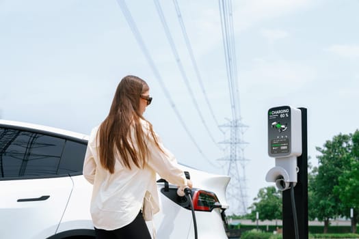 Young woman recharge EV car battery at charging station connected to power grid tower electrical industrial facility as electrical industry for eco friendly vehicle utilization. Expedient