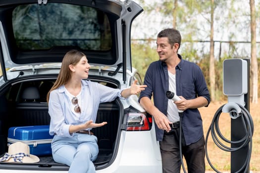 Lovely young couple recharging battery for electric car while relaxing and sitting on the trunk during road trip travel EV car in autumnal forest. Eco friendly travel on vacation during autumn. Exalt