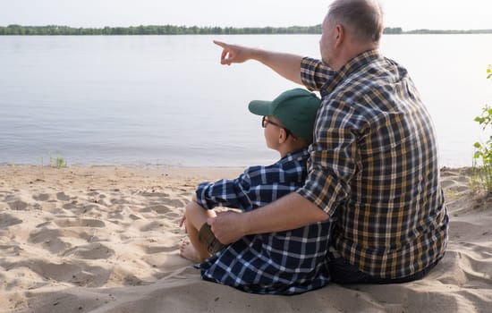 Father's day concept. Rear view of father and son sitting together on the river bank and looking at the water