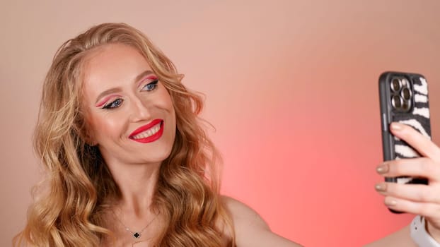 A young woman with red lipstick takes a selfie with long blond hair and makeup. The girl is smiling and looking at the camera close-up