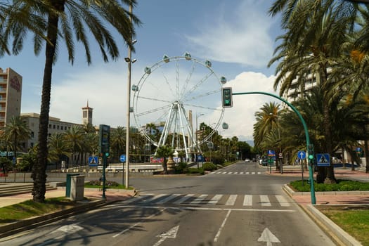 Almeria, Spain - May 25, 2023:A street lined with tall palm trees leading to a towering ferris wheel in the distance, under a clear blue sky.