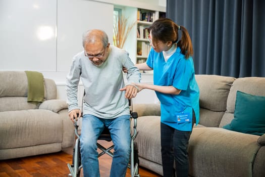 Asian doctor support old man to getting up to exercise, help handicapped elderly stand up, woman nurse assisting helping senior man patient get up from wheelchair for practice walking at home