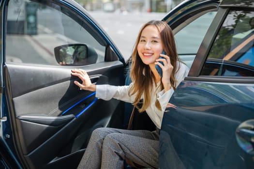 An Asian businesswoman, seated in her luxury car, smiles as she gets a new contract over the phone. Her open car door symbolizes the doorway to business success in the modern city.