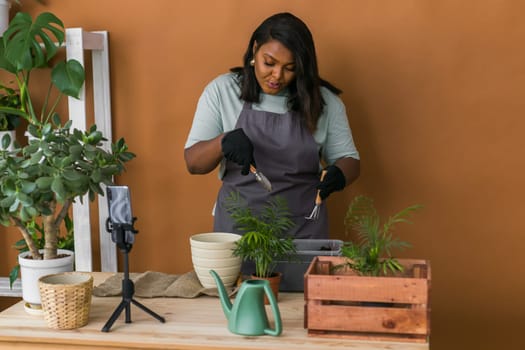 African american girl vlogger influencer or SME owner work on home video camera selfie shoot filming take care home plants and transplanting plant in flowerpot. Home gardening and florist