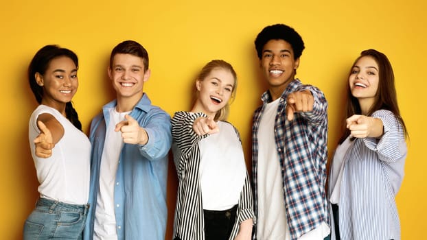 Multiethnic teenagers are standing together, smiling and pointing directly at the viewer. They are casually dressed and appear upbeat. The bright yellow background accentuates the joyful ambiance.