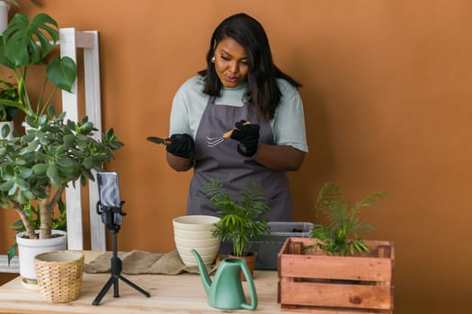 African american girl vlogger influencer or SME owner work on home video camera selfie shoot filming take care home plants and transplanting plant in flowerpot. Home gardening and florist