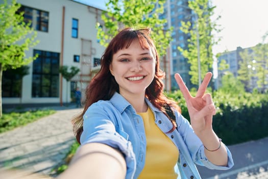 Selfie portrait of young beautiful attractive happy red-haired female looking at camera outdoor. Sunny day teenage girl student 19-20 years old hand showing victory peace gesture educational building