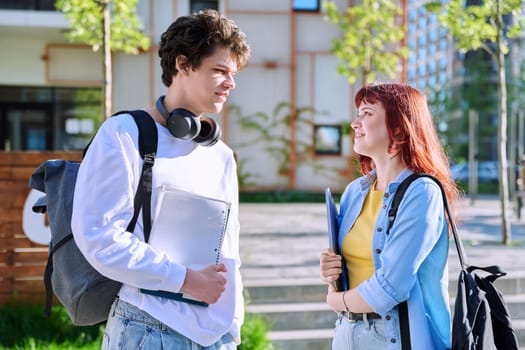 Teenage college students guy and girl talking, standing outdoor near educational building. Youth 19-20 years old, education, lifestyle, friendship concept