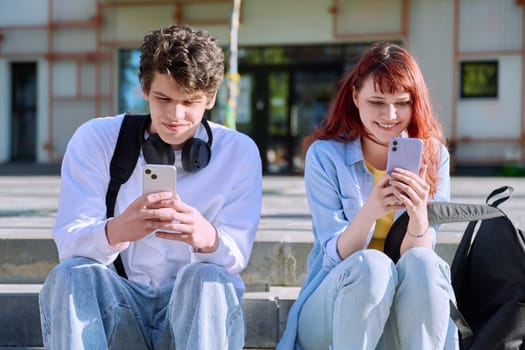 Teenage youth guy and girl university college students sitting outdoor on campus steps using smartphone. Technology, friendship, lifestyle concept