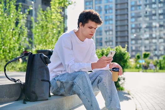 Relaxed resting handsome young male with smartphone, glass of coffee to go, sitting on steps, outdoor. Student 19-20 years old with a backpack, lifestyle, youth concept