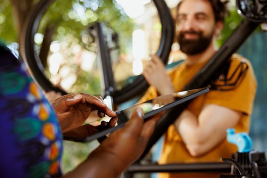 Closeup of black woman grasping phone tablet while enthusiastic young man in background repairs bicycle wheel outdoors. African american woman holding digital device to research fixes for broken bike tire.