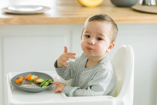 Baby boy sitting in baby chair eating carrot on kitchen background - baby feeding