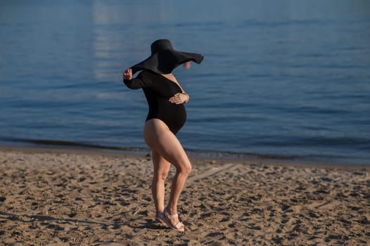 Pregnant woman in a large straw hat and black swimsuit posing on the beach