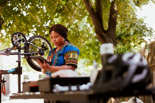 Healthy active female cyclist grasping smart device with instructions for fixing bicycle. Sporty black woman using digital tablet to research bike adjustments for expert maintenance at home yard.