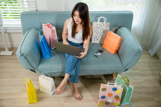 Asian young woman sitting on sofa with shopping bags using laptop computer to online shopping, Happy female satisfied customer with purchase on web site in living room at home