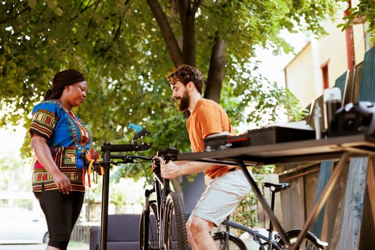 Caucasian man with assistance from african american woman are carefully mounting bicycle to repair-stand for further checking and repairing. Outdoor summer bike maintenance couples hobby activity.