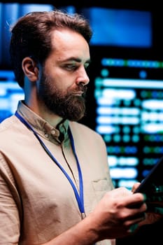 Programmer doing thorough system examination, checking data on tablet of needed debugging in server mainframes housing advanced data storage hardware supporting complex computer operations