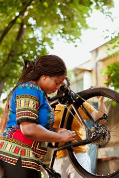 Black woman outdoors checking and fixing bicycle wheel with professional equipments for summer cycling. Young african american woman repairing broken bike tire and chains with tools in yard.