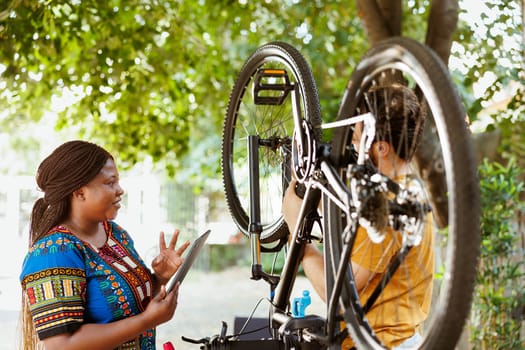 Vibrant black woman happily surfing internet to assist athletic man with bicycle maintenance. Smiling woman with digital tablet gives instructions to active young man to fix bike wheel and chain.