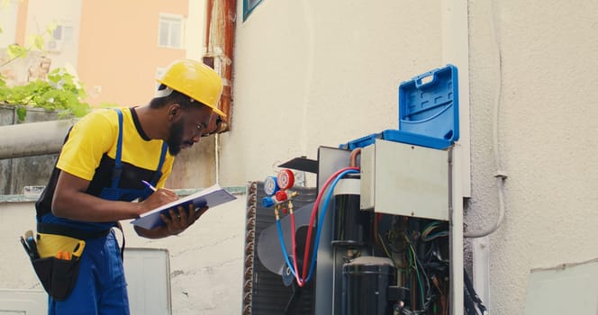 African american electrician doing leak checks and other necessary fixes to prevent major breakdowns. Skillful serviceman verifying air conditioner, writing findings on clipboard