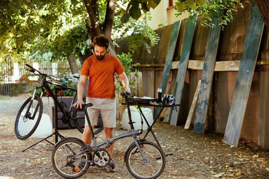 Caucasian male cyclist doing home maintenance in his yard surrounded by collection of professional cycling gear. Sports-loving man ensuring safety of bicycle after repairing.